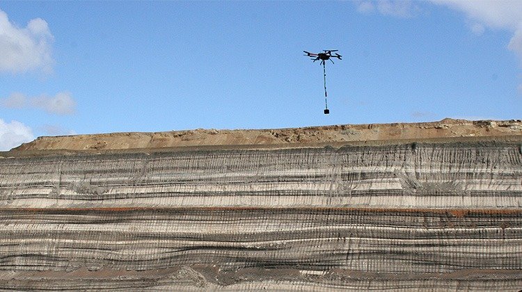 Drone carrying a suspended sensor above exposed geological layers in a quarry.